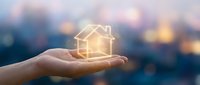 Hand holding glowing house icon with warm light against blurred city background at dusk. Real estate investment and home protection concept for marketing materials.