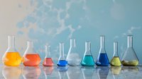 Colorful laboratory flasks and beakers filled with vibrant liquids arranged in a row against a light blue background in a science lab.