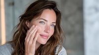 Young woman with blue eyes and brown hair touching her face gently, looking thoughtfully away in soft natural light against blurred background.