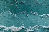Weathered turquoise wall surface with peeling paint and rough texture showing layers of decay, creating an abstract pattern of cracks and flakes.