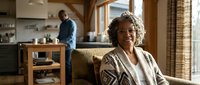 Smiling African American senior woman relaxing on sofa at home while husband cooks in modern kitchen. Mature couple enjoying retirement lifestyle.