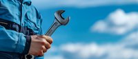 Mechanic holding wrench against blue sky with white clouds, close-up view of hands in blue uniform. Professional maintenance and repair service concept.