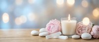 Spa wellness scene with lit candle, pink peonies and white stones on wooden surface with soft bokeh lights creating peaceful relaxation atmosphere.