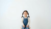 Young Asian girl with braided hair wearing denim overalls and cream long-sleeve shirt, smiling at camera against white background, suitable for children's fashion and lifestyle content.