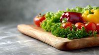 Fresh colorful vegetables including cherry tomatoes, lettuce, parsley and bell peppers arranged on wooden cutting board against gray background.