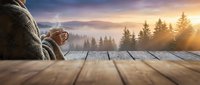 Person in cozy sweater holding warm drink on wooden deck overlooking misty forest landscape at golden sunrise. Peaceful morning relaxation concept.