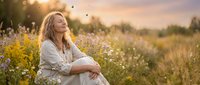 Pregnant woman in white dress sitting peacefully in wildflower meadow during golden hour sunset with warm natural lighting and serene countryside atmosphere.