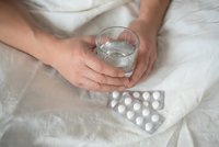 Person holding glass of water with medication pills on bed for health treatment and wellness routine.