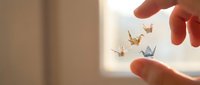 Hand holding delicate origami paper cranes suspended in air against soft natural light, symbolizing peace, wishes, and mindfulness for wellness and meditation concepts.