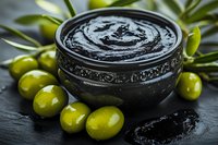 Black cosmetic charcoal face mask in vintage ceramic bowl surrounded by fresh green olives and olive leaves on dark stone background. Natural skincare concept.