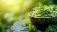 Fresh homemade guacamole in rustic stone bowl garnished with mint leaves, served on dark slate against blurred green garden background with sunlight.