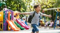 Happy young Asian boy with backpack running with arms outstretched at colorful playground. Joyful child enjoying outdoor recreation and active play time.