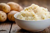 Creamy mashed potatoes with butter and spices in white bowl, fresh raw potatoes on rustic wooden table background. Traditional comfort food side dish.