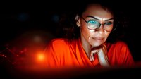 Young African American woman in orange attire and stylish glasses illuminated by warm glowing lights, hand resting on chin, expressing thoughtful contemplation against dark background.