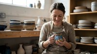 Smiling Caucasian woman artisan in apron holding handmade ceramic mug in pottery studio with shelves of handcrafted dishes.