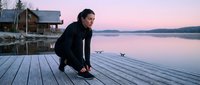 Man in black jacket crouching on wooden dock at sunrise, gazing at calm lake water with cabin and mountains in background. Contemplative outdoor lifestyle scene.