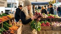 Young blonde woman in beige apron and knit hat selecting fresh radishes at bustling farmers market with colorful organic vegetables and produce stands.