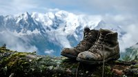Worn hiking boots on mossy rock with snow-capped mountain peaks in background. Adventure trekking footwear after long journey through alpine wilderness terrain.