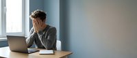 Stressed young businessman holding head in hands at office desk with laptop showing work burnout and mental health challenges in corporate environment.