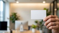 Person of South Asian descent holding blank white business card in modern office environment with plants and natural lighting, suitable for corporate branding and professional networking concepts.