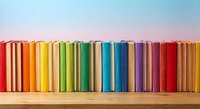 Colorful books arranged in rainbow spectrum on wooden shelf against pastel sky background for education and reading concepts.