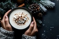 Hands holding warm winter coffee with cocoa powder and cream surrounded by festive pine branches and cones on dark background for cozy holiday moments.