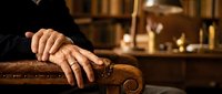 Senior man's weathered hands resting on ornate wooden chair armrest in warm library setting with books and soft lighting for retirement concepts.