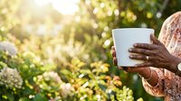 Woman with dark skin holding white mug in garden with blooming flowers and sunlight, wearing patterned clothing, outdoor beverage lifestyle scene.