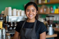 Young Asian female barista wearing black apron smiling warmly at camera in coffee shop setting with coffee equipment and supplies visible in background. Professional service industry portrait.