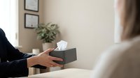 Professional counselor offering tissue box to client during therapy session in modern office setting with certificates on wall.