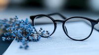 Black-rimmed eyeglasses resting on open book pages with delicate blue dried flowers creating elegant still life composition for reading and literature concepts.