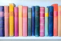 Row of vintage hardcover books in various bright colors including pink, yellow, blue, and green arranged on white shelf, spines showing classic binding details.