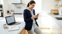 Woman in casual business attire working from home kitchen, using smartphone while laptop and notebook sit on marble counter, multitasking with technology and domestic space.