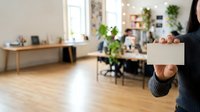 Businesswoman holding blank cardboard box in modern office workspace with wooden furniture, natural light, and green plants, suitable for relocation, moving services, and corporate business concepts.