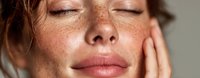 Young woman with freckles touching face with closed eyes in natural skincare routine. Gentle self-care moment showing healthy glowing skin and natural beauty.