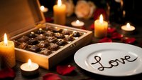 Romantic candlelit table with luxury chocolate box and white plate with Love written in chocolate, rose petals, warm candles for Valentine's Day.