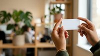 Person holding blank white business card in modern office environment with plants and colleagues in background.