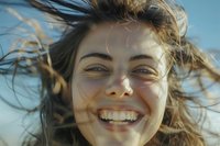 Young Caucasian woman with windblown hair laughing joyfully outdoors in warm sunlight, showing genuine happiness and natural beauty in closeup portrait.