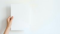 Hand holding blank white paper sheet against neutral wall background, suitable for mockups, presentations, and advertising campaigns.
