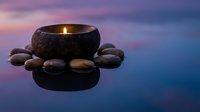 Stone bowl candle with smooth river rocks on reflective water surface during twilight hour creating peaceful spa atmosphere for meditation and relaxation.