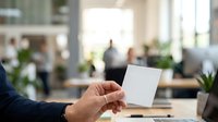 Male hand holding blank white card in modern office environment with blurred colleagues and workspace in background for business branding mockup.