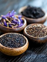 Wooden bowls with black tea leaves, chia seeds, dried purple flowers and black salt on dark wooden surface, macro food photography for wellness and tea culture.