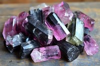 Raw tourmaline crystals in pink and black colors on wooden surface, showing natural mineral formation with translucent and opaque specimens in varying sizes.
