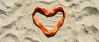 Orange heart-shaped object resting on textured sand surface with natural shadows and footprint impressions creating romantic beach scene for summer love concepts.