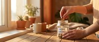 Woman putting coins into glass jar labeled home savings on wooden table with coffee cup and plants in warm sunlit kitchen for financial planning goals.