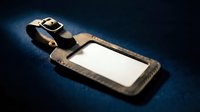 Black leather luggage tag with blank white label and metal buckle on dark blue surface for travel identification and personalization.