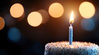 Single lit blue striped birthday candle on white frosted cake with sprinkles against dark background with soft glowing bokeh lights creating festive atmosphere.