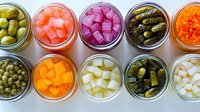 Colorful assortment of pickled vegetables and garnishes in glass jars viewed from above, including olives, onions, carrots, and cornichons arranged in neat rows.