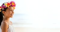 Young girl with long dark hair wearing colorful flower crown at beach, smiling toward ocean horizon in white sleeveless dress.