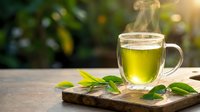 Hot green tea steaming in glass mug on wooden board with fresh leaves against blurred natural background. Healthy herbal beverage for wellness and relaxation.